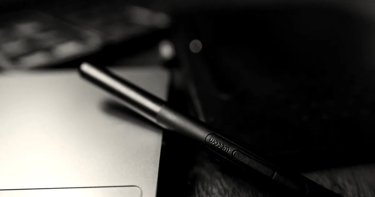 A black Wacom stylus pen resting on a laptop next to a graphics drawing tablet on a wooden desk.