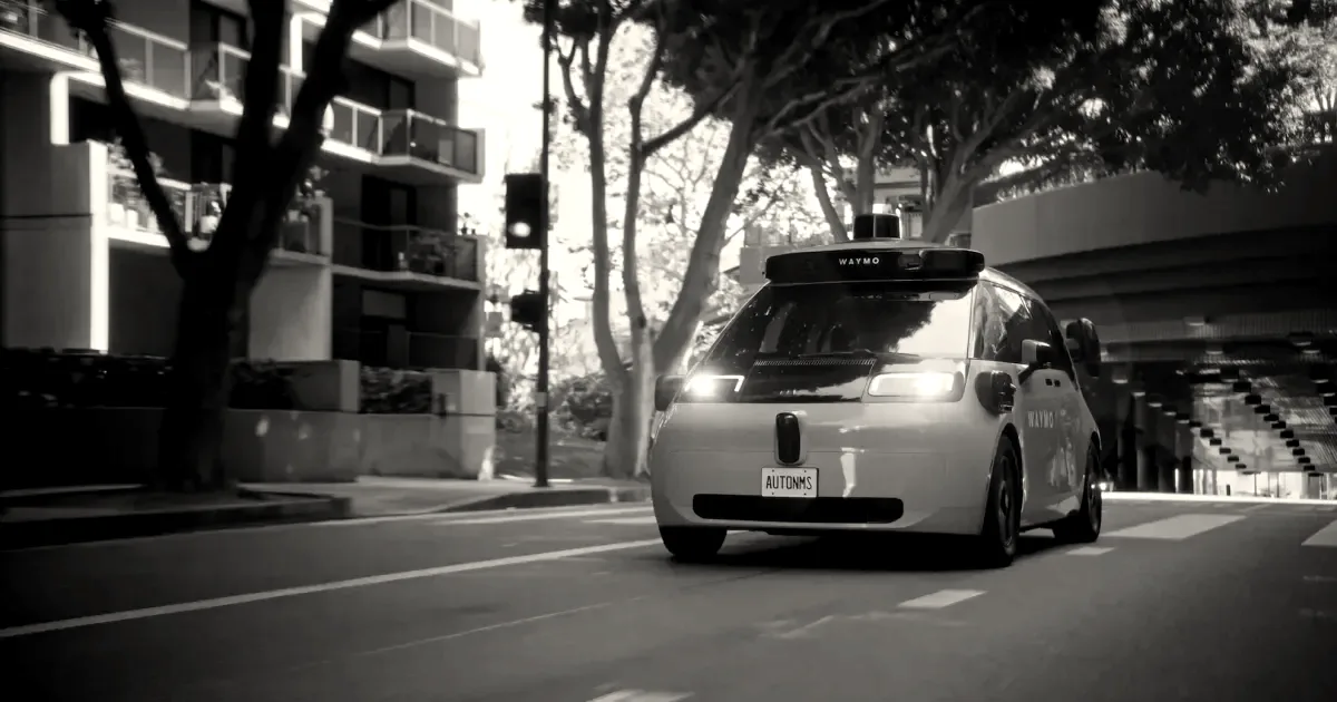 A light blue autonomous Waymo vehicle driving on an urban city street with headlights illuminated.