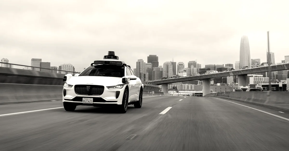A white Waymo autonomous Jaguar SUV driving on a multi-lane freeway with a city skyline in the background.