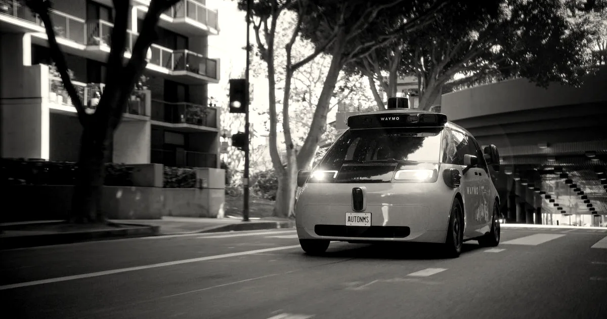 A light blue autonomous Waymo vehicle driving along a city street lined with trees and modern buildings.