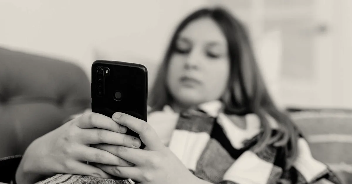 A teenage girl lying down on a couch while holding and using a black smartphone.