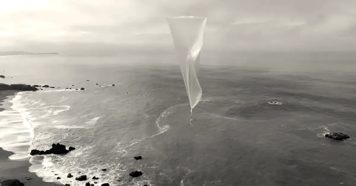 A translucent high-altitude weather balloon ascends over a coastline with ocean and rocky shore below.