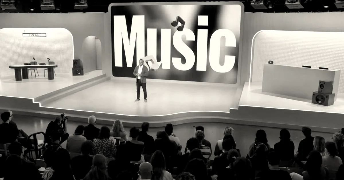 A presenter stands on stage before an audience in front of a giant screen displaying YouTube Music branding.