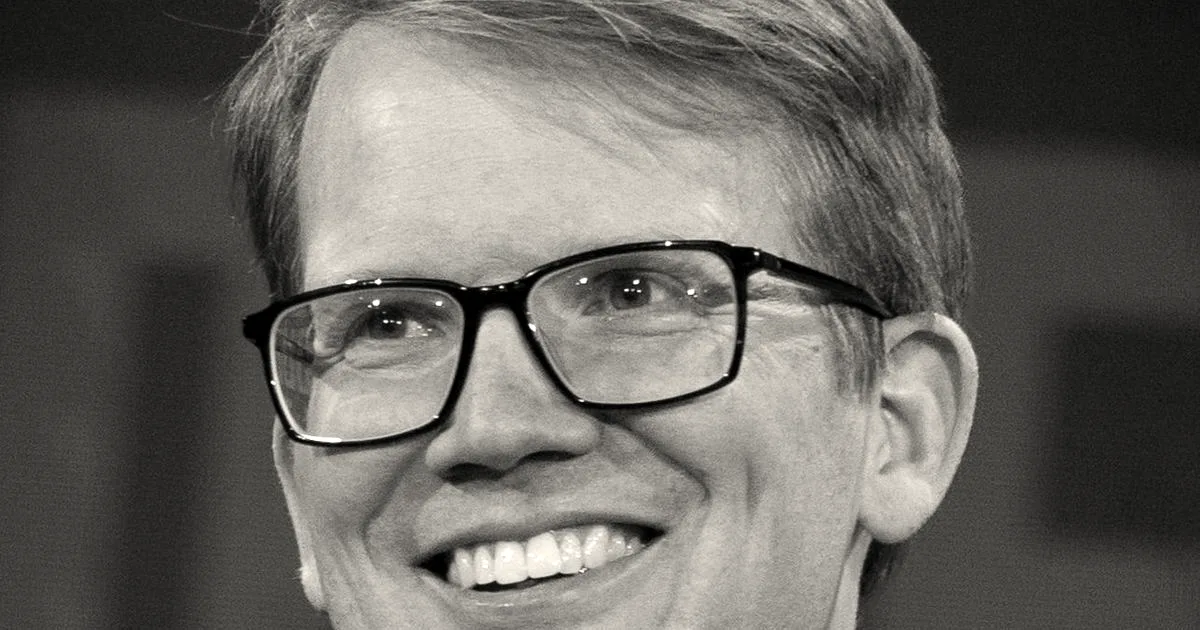 A close-up headshot of Hank Green smiling and holding a microphone while wearing glasses.