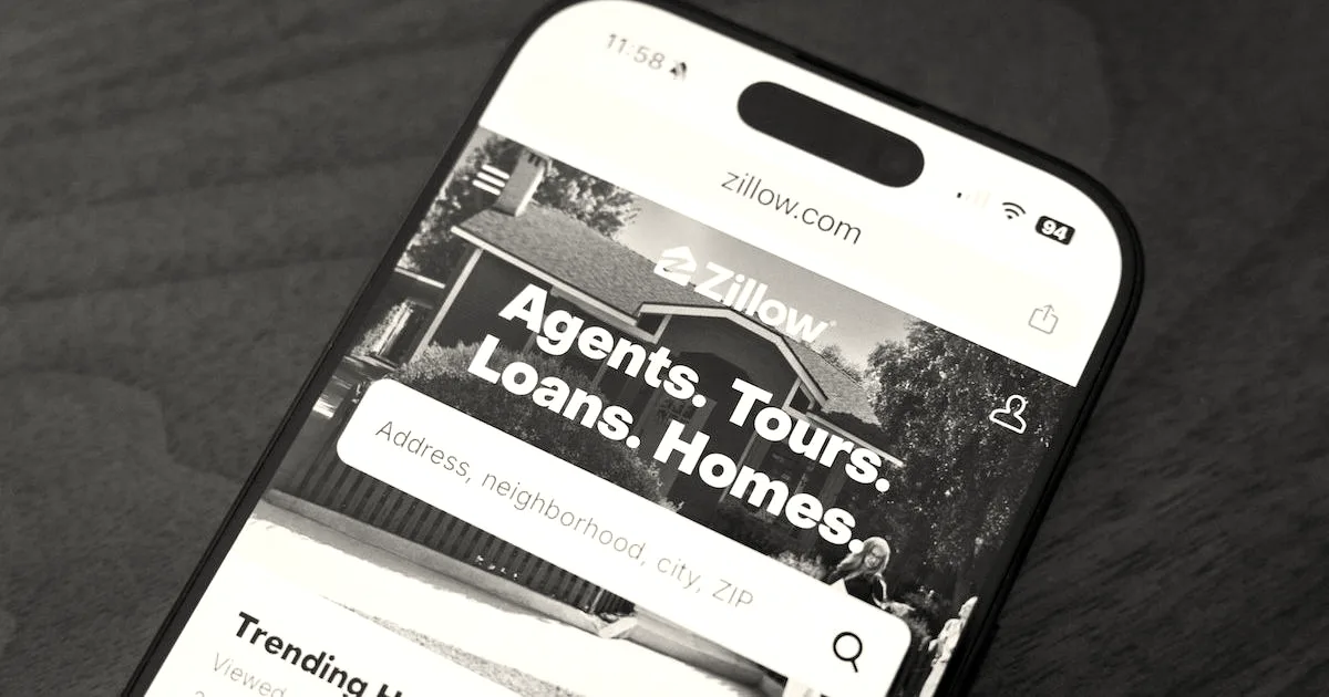 A smartphone resting on a wooden surface displays the Zillow website homepage on its screen.