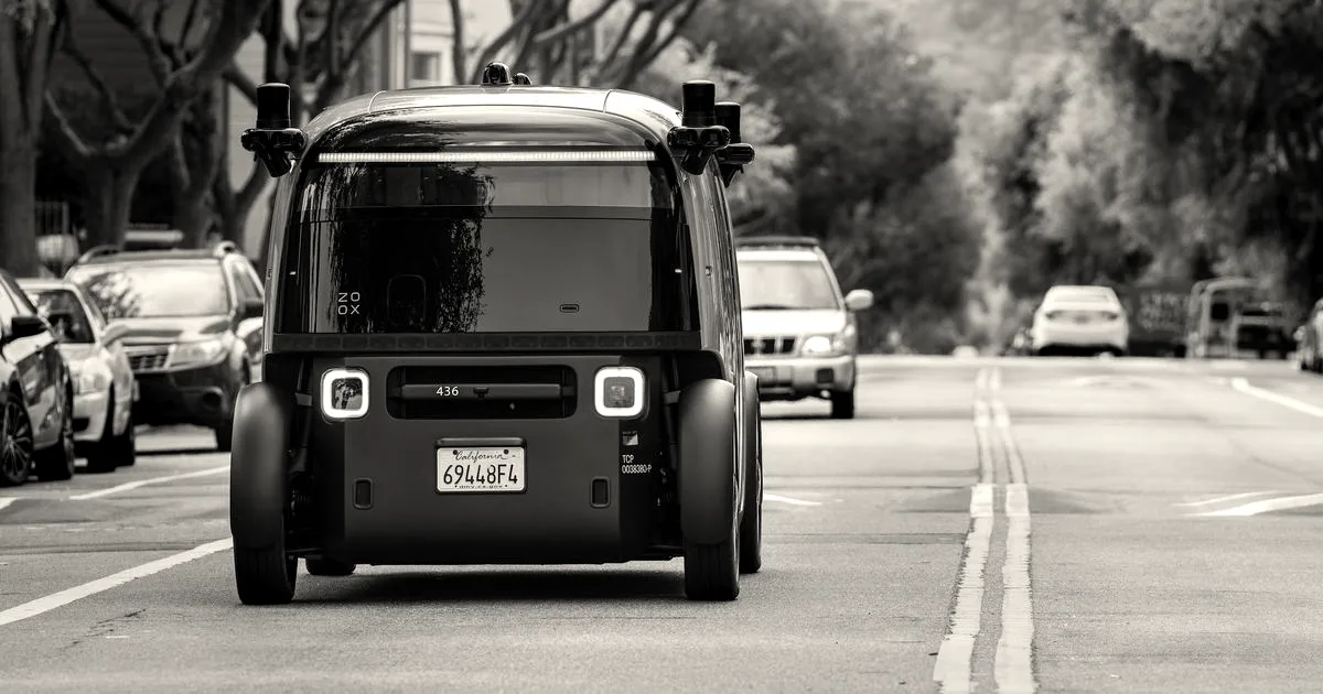 Rear view of a Zoox autonomous robotaxi driving down a city street with other vehicles nearby.